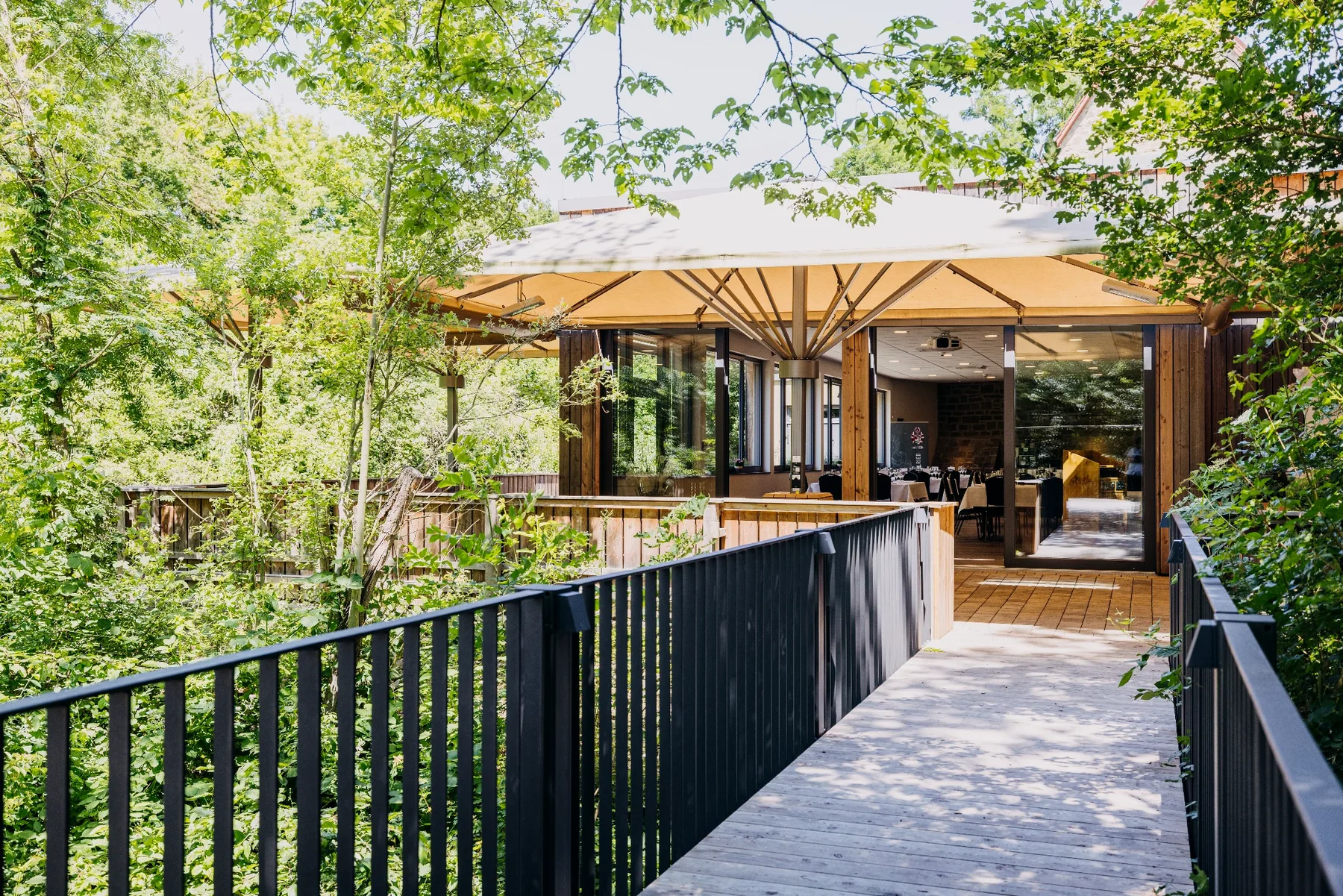 Holzsteg mit Geländer als Zugang zur überdachten Terrasse des Ulrichsaals auf Gut Wöllried, umgeben von Bäumen